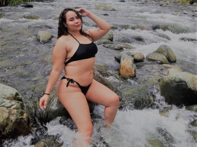EveNailah with curly brown hair, black bikini, medium tits, curvy ass, tattoo on right shoulder, kneeling in rocky stream, water splashing, smiling, right hand in hair.