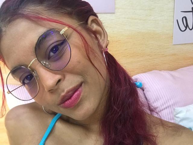 HilaryCook tiene lápiz labial rosa, sombra de ojos morada y cabello rojo-rosado recogido en una coleta. Lleva gafas redondas y una correa azul, con una sonrisa sutil. Sus pechos son pequeños..