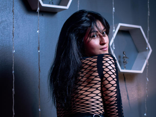 JessiLemus, con cabello negro largo, mirando por encima del hombro, vestida con una blusa de malla negra, que deja ver su espalda y sus senos, frente a una pared oscura con estantes hexagonales..