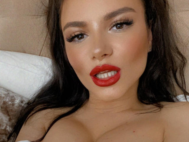 LexaKiss has red lipstick, dark brown eyes, long eyelashes, and glossy skin. She's looking at the camera with slightly parted red lips, showing her teeth. Boobs visible.