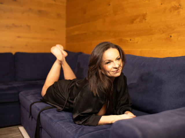 HelenDesire, allongée sur un canapé bleu, vêtue d'une robe noire, avec ses fesses levées et les pieds croisés, sourit à la caméra. Ses seins sont partiellement visibles..