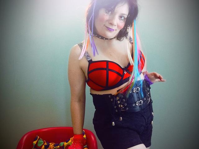 EverlyRays with colorful hair, red sports bra showing her tits, black lace-up corset, black shorts, red gloves, sitting on red chair, smiling, wearing choker, against plain background.