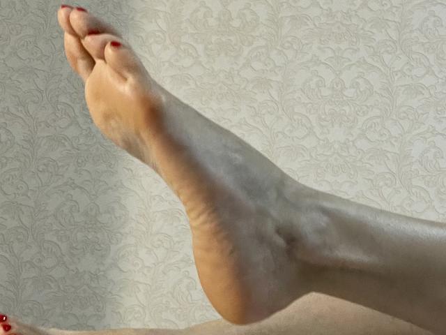 TheHottestBlonde's bare foot is raised with red-painted toenails, showing smooth, tan skin and slight heel crease against a patterned wallpaper.