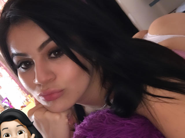 ShantallParis with black hair, full pink lips, and brown eyes, pouting, lying on a purple pillow, wearing a white top, showing her ass in the background.