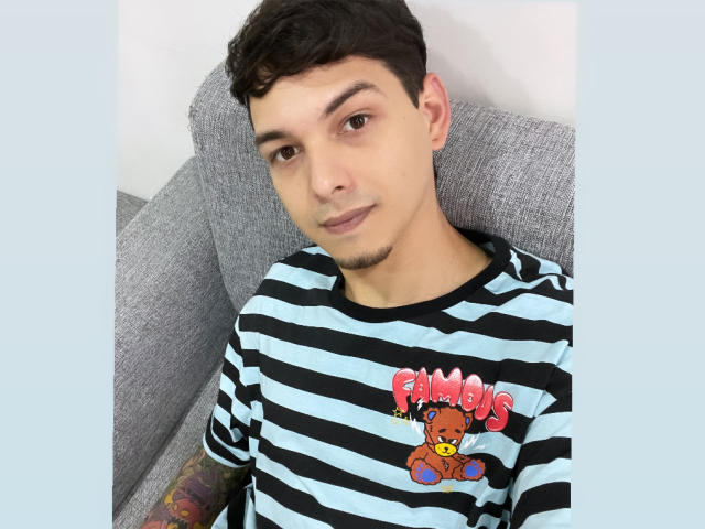Elyan with light skin, short curly brown hair, small goatee, black striped shirt with "Pumbas" teddy bear, tattoo on left arm, lying on gray couch, slight smirk.