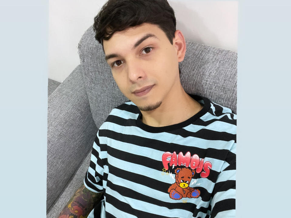 Elyan with light skin, short curly brown hair, small goatee, black striped shirt with "Pumbas" teddy bear, tattoo on left arm, lying on gray couch, slight smirk.