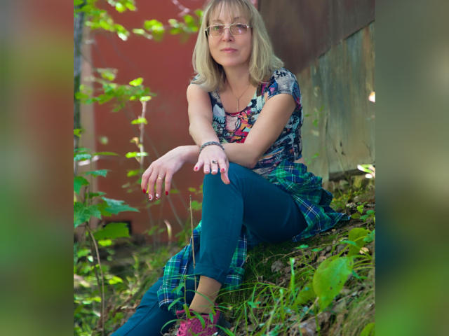 IrunchaArt with blonde hair, glasses, floral top, blue plaid skirt, green pants, sitting on grass, hands on knee, red nail polish, wooden wall in background, subtle smile.