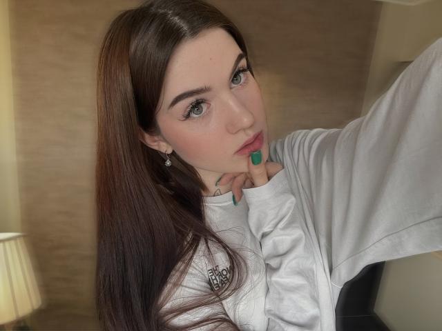 Nicole, con lunghi capelli castani, occhi verdi e unghie verdi, indossa una felpa grigia, si tiene il mento e guarda la fotocamera con un sorriso appena accennato..