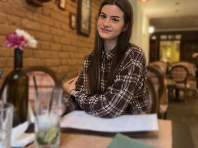 Nicole, mit langen braunen Haaren, trägt ein kariertes Hemd, sitzt an einem Tisch, lächelt, hat die Arme verschränkt, im Vordergrund befinden sich eine Glaswasserflasche, eine Blume und Papier..