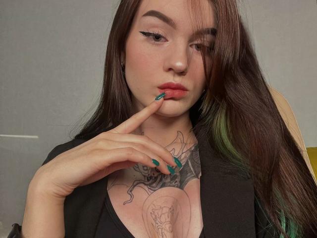 Nicole, avec ses longs cheveux bruns, son blazer noir et ses ongles vert turquoise, touchant ses lèvres rouges avec son doigt, montrant un tatouage sur la poitrine et des mèches vertes dans ses cheveux..