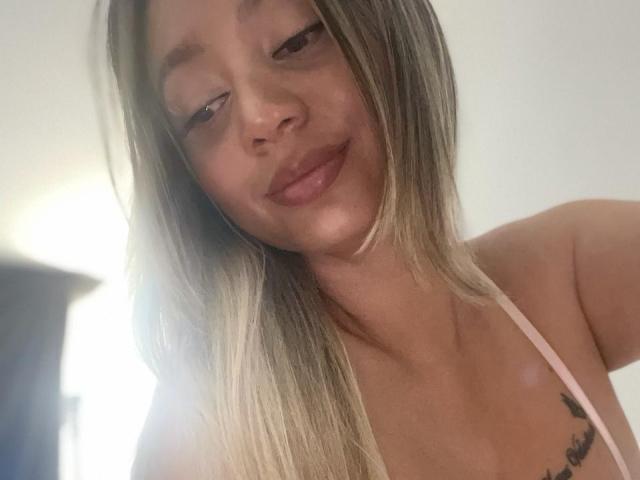 Sophie Lombardo mit langen blonden Haaren, einem pinken Haltertop und einem kleinen Tattoo auf der rechten Brustseite, lächelt sanft in die Kamera, hat braune Augen und helle Haut..