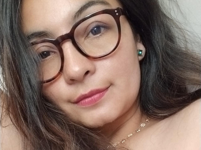 A CandySweetDee usa óculos marrons, tem cabelo ondulado castanho escuro e um tom de pele marrom claro, com um sorriso discreto, usando batom rosa e um brinco verde, mostrando seus seios..