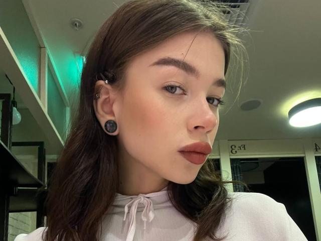 TuneGodness a un joli visage, des cheveux bruns ondulés, de petits bouchons d'oreille noirs et un piercing au septum, et elle porte une chemise blanche avec un léger rougissement sur ses joues..