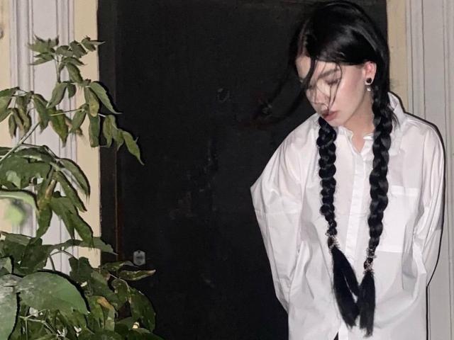 TuneGodness with black braids, wearing a white shirt, hands in pockets, looking down, dark door in background, green leafy plant on left.