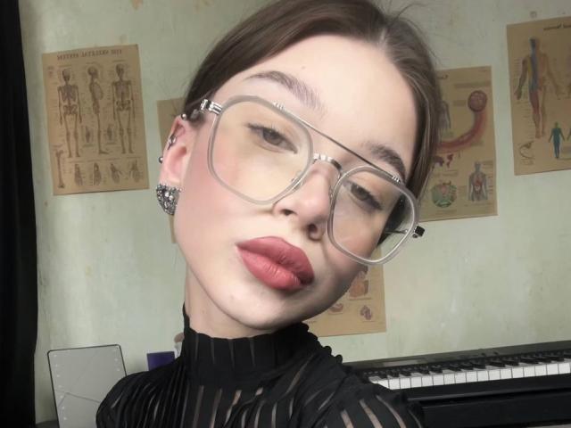 TuneGodness, avec des lunettes, du rouge à lèvres et un anneau de nez, portant un haut noir transparent, mettant en valeur un piercing septum et des élargisseurs d'oreille, regardant la caméra..