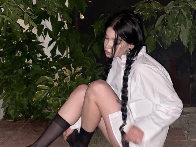 TuneGodness has long black braids, wears a white shirt, black stockings, and is crouching with knees up, showing pale skin and a serious face at night.