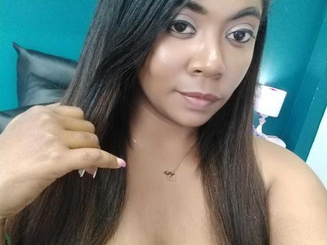 Nathy Martinez, met lang bruin haar, houdt een sleutel vast, glimlacht, draagt een hartvormige ketting, heeft roze nagels, een gebruinde huid en subtiele make-up, en laat haar kleine borsten en schattige gezicht zien..