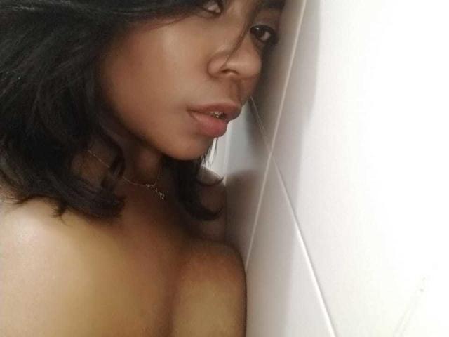 NathyMartinez has dark brown wavy hair, medium brown skin, and a lip piercing, with bare shoulders and a small necklace, leaning against a white wall.