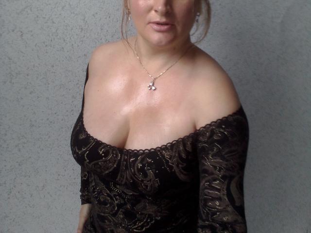 KelliRedhaer, una donna corpulenta con un seno abbondante, indossa un abito nero in pizzo con una spalla scoperta, una collana con un piccolo ciondolo e i capelli biondi raccolti in uno chignon morbido, sullo sfondo di un muro grigio..