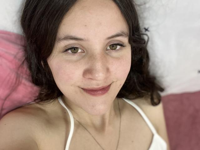 ChloeDulce hat hellbraune Augen, dunkelbraunes, gewelltes Haar und ein zartes Lächeln. Sie trägt ein weißes Top mit Halter und im Hintergrund sind rosa und weiße Kissen zu sehen..