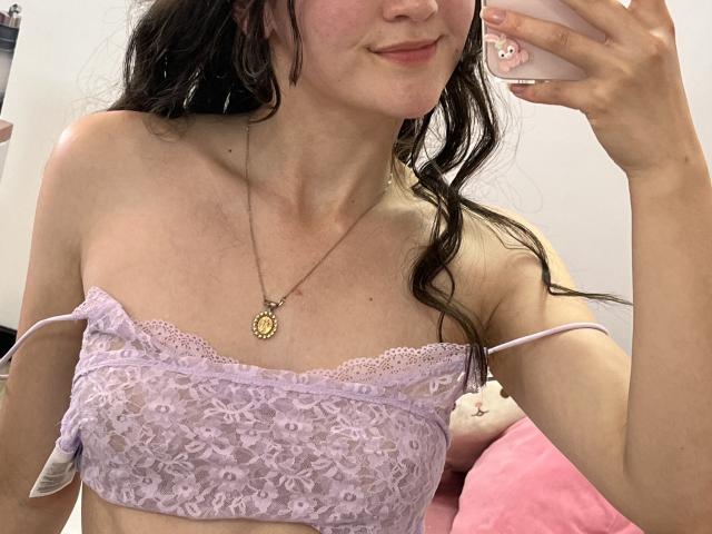 ChloeDulce, com cabelos castanhos ondulados, vestindo um sutiã de renda lilás, mostrando seios pequenos, segurando um telefone, sorrindo, com um colar de ouro, uma cama rosa ao fundo..
