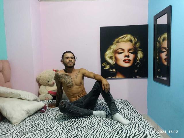 shirtless JeiLerth with tattoos, dark curly hair, and beard, sitting on zebra-pattern bed, black ripped jeans, white socks, next to beige teddy, Marilyn Monroe painting.