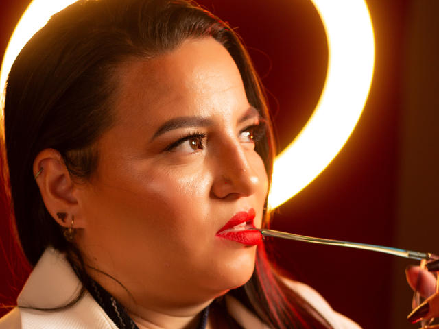SkayLaraWalker's face with glossy red lips, holding a lipstick with a reflective circular light behind her. She has dark hair, brown eyes, and a white coat.