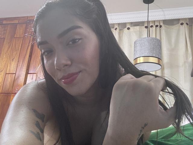 SharonKlart with dark hair, holding a lock, showing a tattoo on her arm, red lipstick, and a cute smile, in a selfie with wood-paneled wall and lamp.