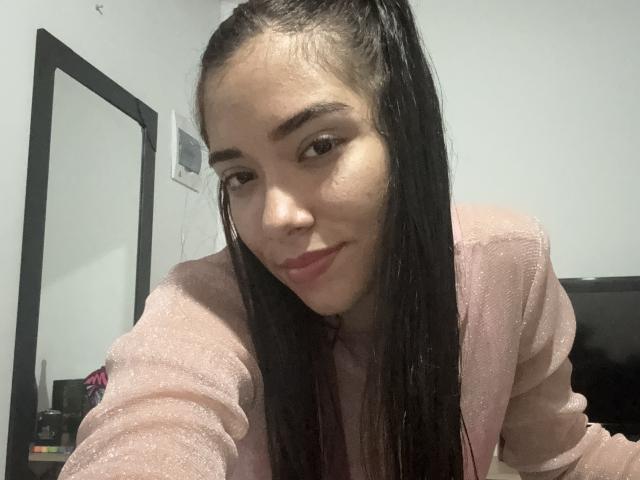 SharonKlart with long black hair in a ponytail, wearing a pink sparkly top, smiling with pink lipstick, brown eyes, and light skin. Her ass is slightly visible.