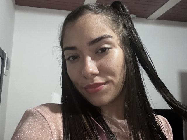 SharonKlart with long black hair in pigtails, wearing a sparkly pink top, smiling slightly, showing a cute face with light skin and subtle makeup.