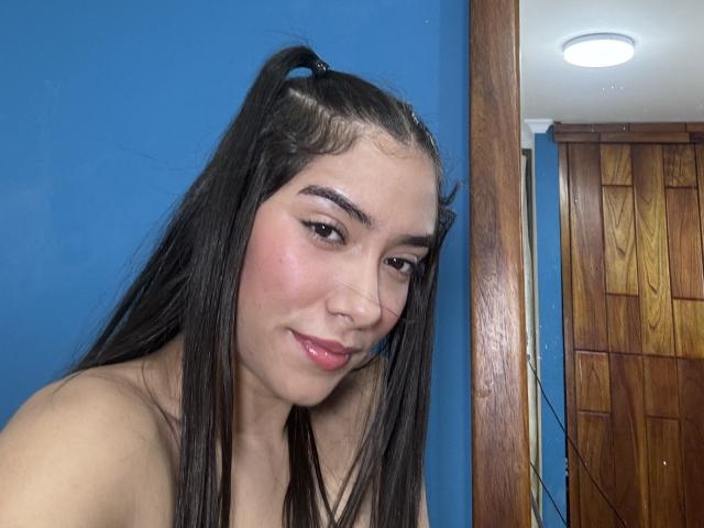 SharonKlart has long black hair in pigtails, pink lipstick, and a nose ring, smiling with pink cheeks, in a blue-walled room with wooden doors in the background.