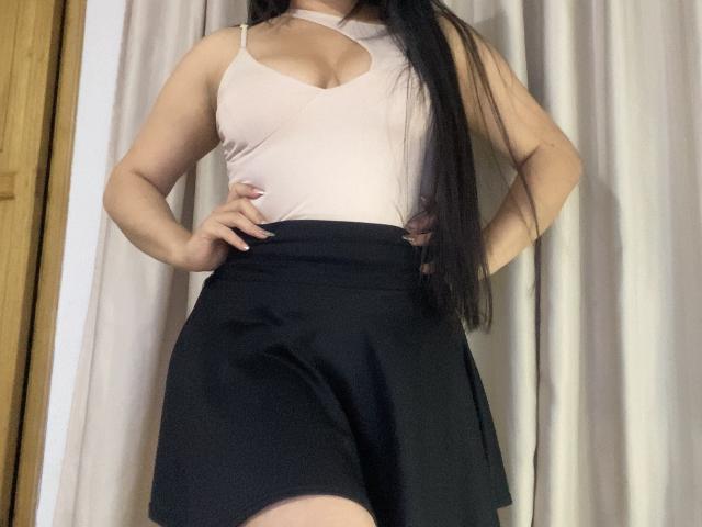 MiaDuboisX with long black hair, pale pink tank top showing her tits, black skirt, hands on hips, standing in a room, curtains in background.