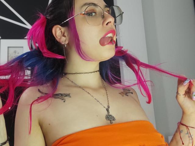NatashaLau with pink and blue hair, glasses, tongue out, orange strapless top, tattoos on shoulders, necklace, holding pink hair, pale skin, earrings, looking at camera, slightly open mouth.