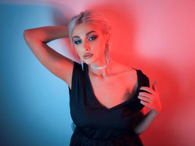 KylieJonnes mit blonden Haaren, blauen Augen und Smokey-Eyes-Make-up, trägt ein schwarzes Kleid, eine Halskette und Ohrringe, eine Hand im Haar, rotes und blaues Licht, sexy, selbstbewusst, blickt in die Kamera..