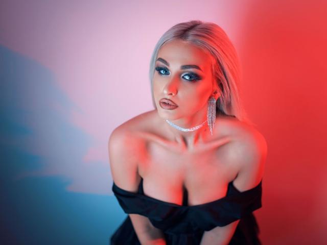 KylieJonnes with platinum blonde hair, blue eye makeup, and a black off-shoulder dress, showing her big tits, wearing a sparkly choker and diamond earrings, in red-blue lighting.