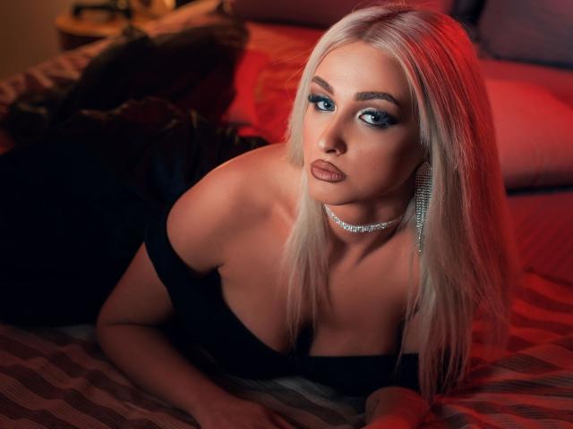 KylieJonnes with platinum blonde hair, blue eyes, and a diamond choker, lying on a bed, wearing an off-shoulder black dress, showcasing her big tits and pouty lips.