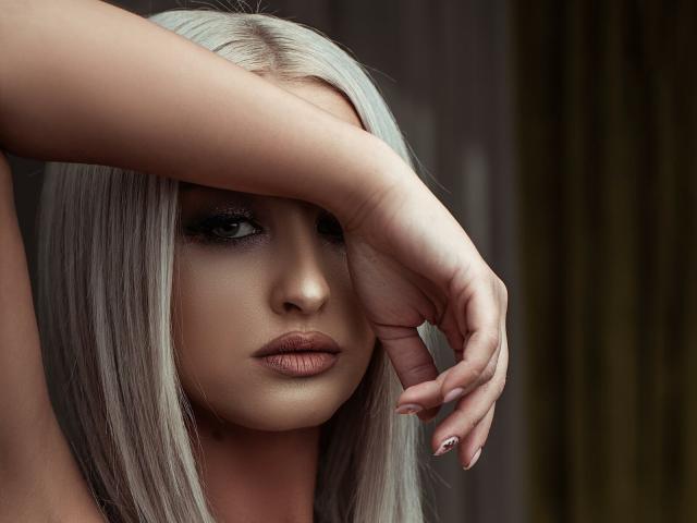 KylieJonnes, con su largo cabello plateado, se cubre los ojos con la mano derecha, tiene labios carnosos y fruncidos y una expresión seria. Sus uñas cuidadas son visibles..