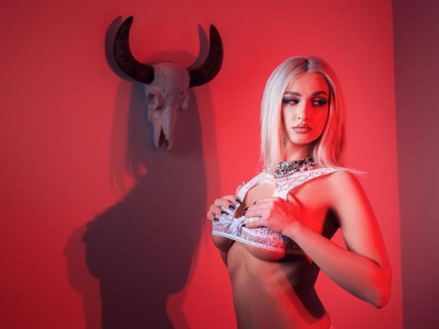 KylieJonnes, con i capelli biondi, indossa pizzo, tiene le mani sui seni, di fronte a un teschio con corna, luce rossa, ombra sul muro, guarda in basso, seducente, sexy..