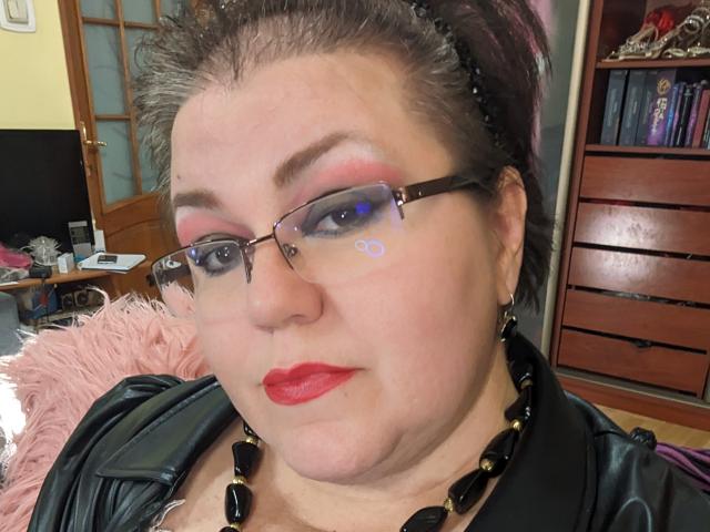 KaryQueen with red lipstick, black glasses, and black beaded necklace, wearing a leather jacket, has short spiky brown hair, and a pink fluffy chair in the background.