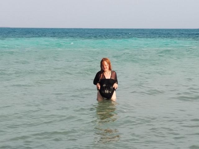 VioletaMora with red hair, black wet shirt, and black bikini bottom, standing in clear blue-green water, holding a black bag, her tits partially visible above water.