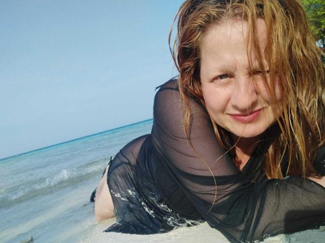 VioletaMora with wet, blonde hair, wearing a sheer black top, lying on a beach, smiling, showing her tits, and her ass, with ocean waves in the background.