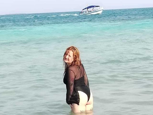VioletaMora with wet, red hair, wearing a black mesh top and white butt-puffy, kneeling in shallow water, looking back at the camera, with a boat in the background.