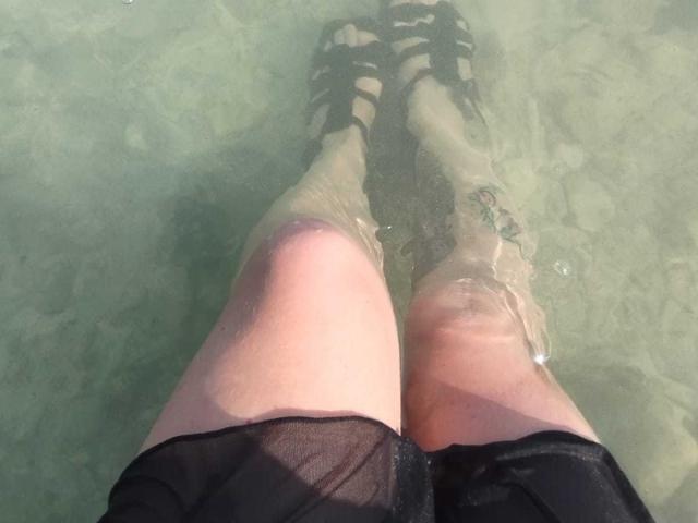 VioletaMora's legs in water, wearing black strappy sandals, black sheer skirt, visible tattoo on right thigh, knees slightly bent, water level just above ankles.