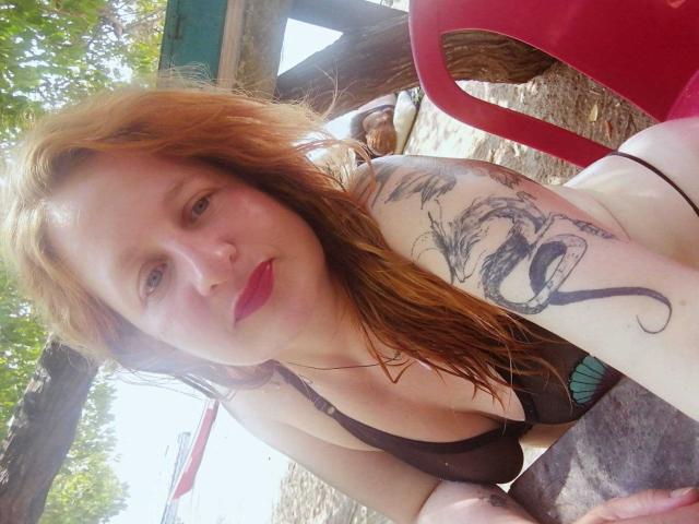 VioletaMora with red hair, pale skin, and tattoos on her arm, wearing a black bikini, has red lipstick and is leaning forward with a red chair behind her.