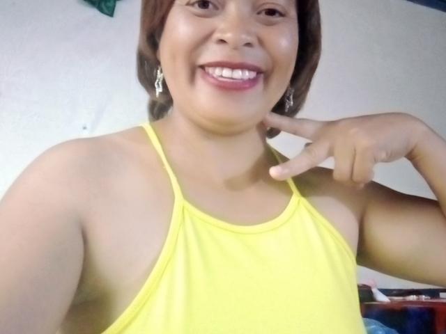 Rahel69 with medium brown skin, smiling, wearing a yellow tank top, pointing at her chin with her right index finger, has short brown hair and small earrings.