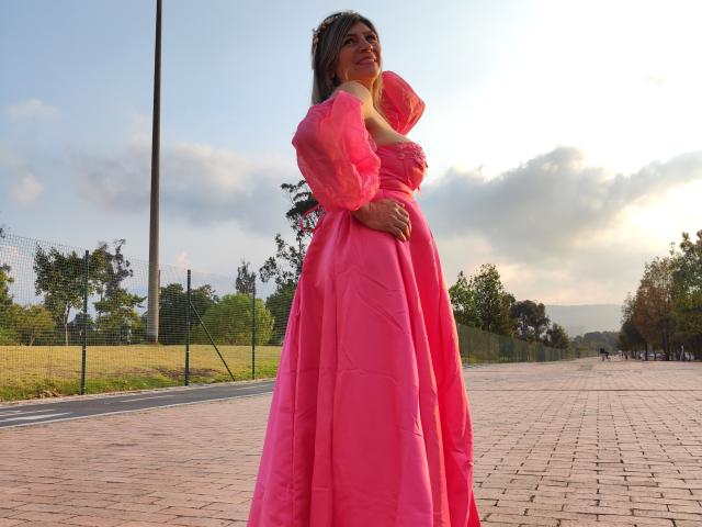 Celeste Lincon, vestida con un vestido de bola rosa con mangas abullonadas, está de pie en un camino de ladrillos, con una mano en el pecho, mirando hacia arriba, y sus pechos son ligeramente visibles debajo del vestido..
