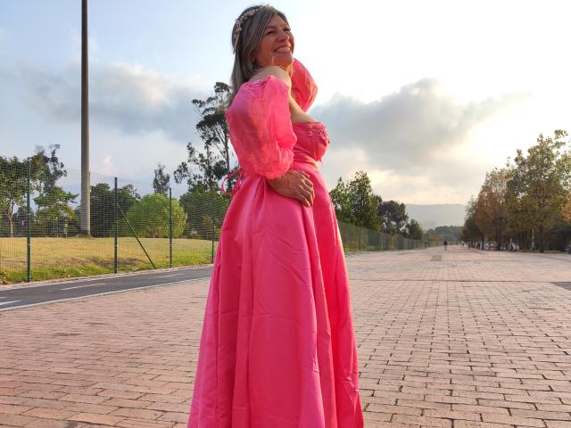 CelesteLincon in a pink ball gown, standing with her hand on her ass, smiling, and looking over her shoulder, on a brick pathway, with trees in the background.
