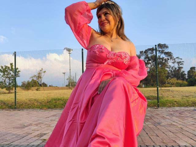 Celeste Lincon, con un vestido rosa de corte asimétrico con encaje, sentada con una mano en la cabeza, mostrando sus senos, sonriendo, sobre una superficie de ladrillo, con un cielo azul..