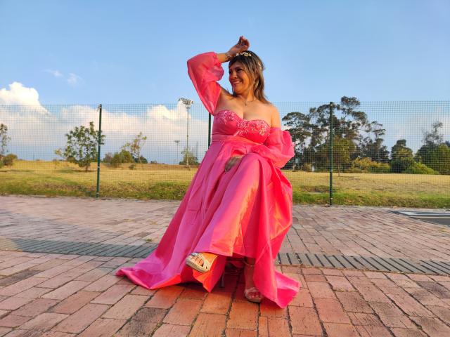 CelesteLincon with pink dress, tits visible under sheer fabric, smiling, arm raised, standing on brick patio, clear blue sky, green field, fence in background, tan sandals.