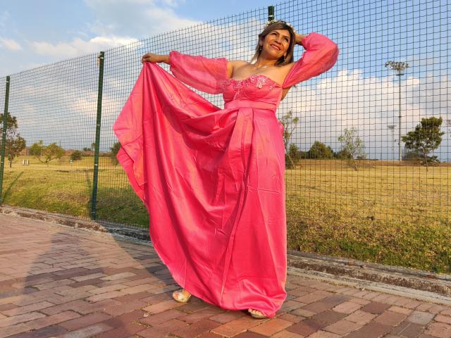 CelesteLincon stands in pink satin dress, holding skirt, showing tits in a mesh fence background, smiling, hand in hair, pink dress, yellow sandals, brick path, sunny day.